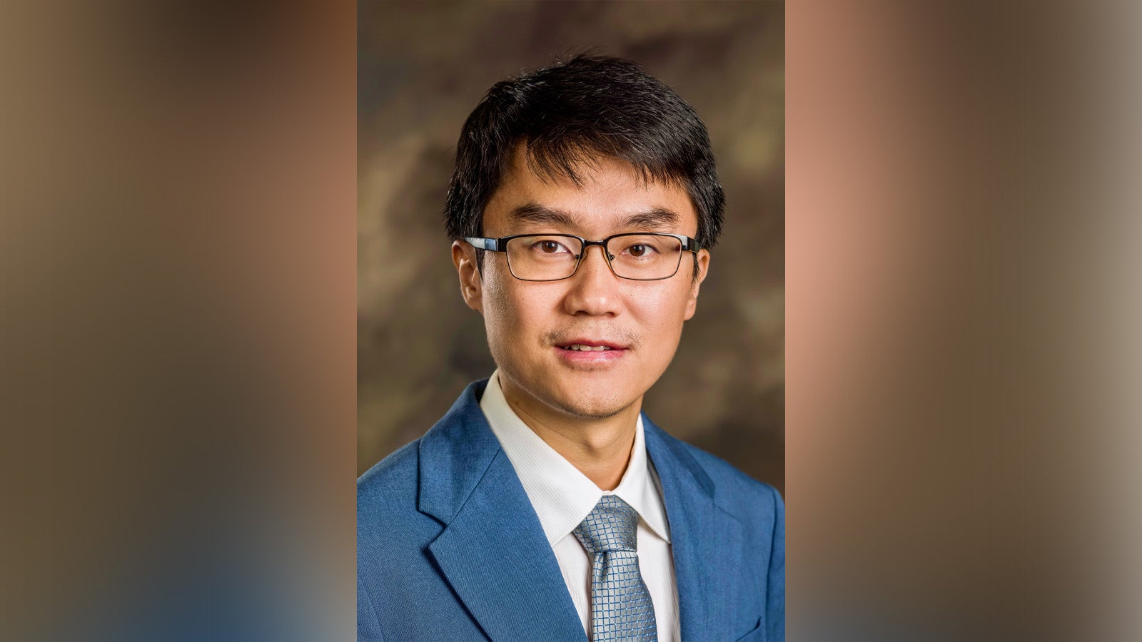 Portrait of an Asian man in a blue suit and tie, wearing glasses, looking at the camera with a neutral expression against a blurred brown backdrop.