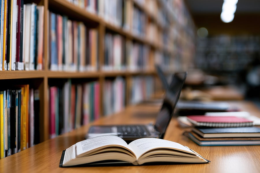 Open book spread on a wooden desk in a library, with a laptop and notebooks nearby.
