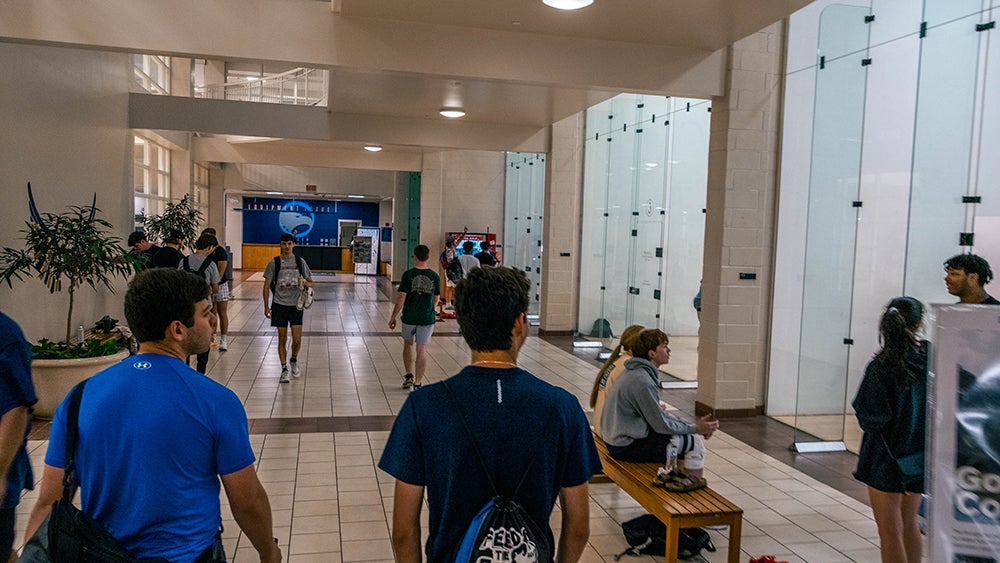 Busy indoor campus hallway with students walking; glass-walled rooms on the right and a person seated on a wooden bench on the right side out of focus shows a modern atrium feel.