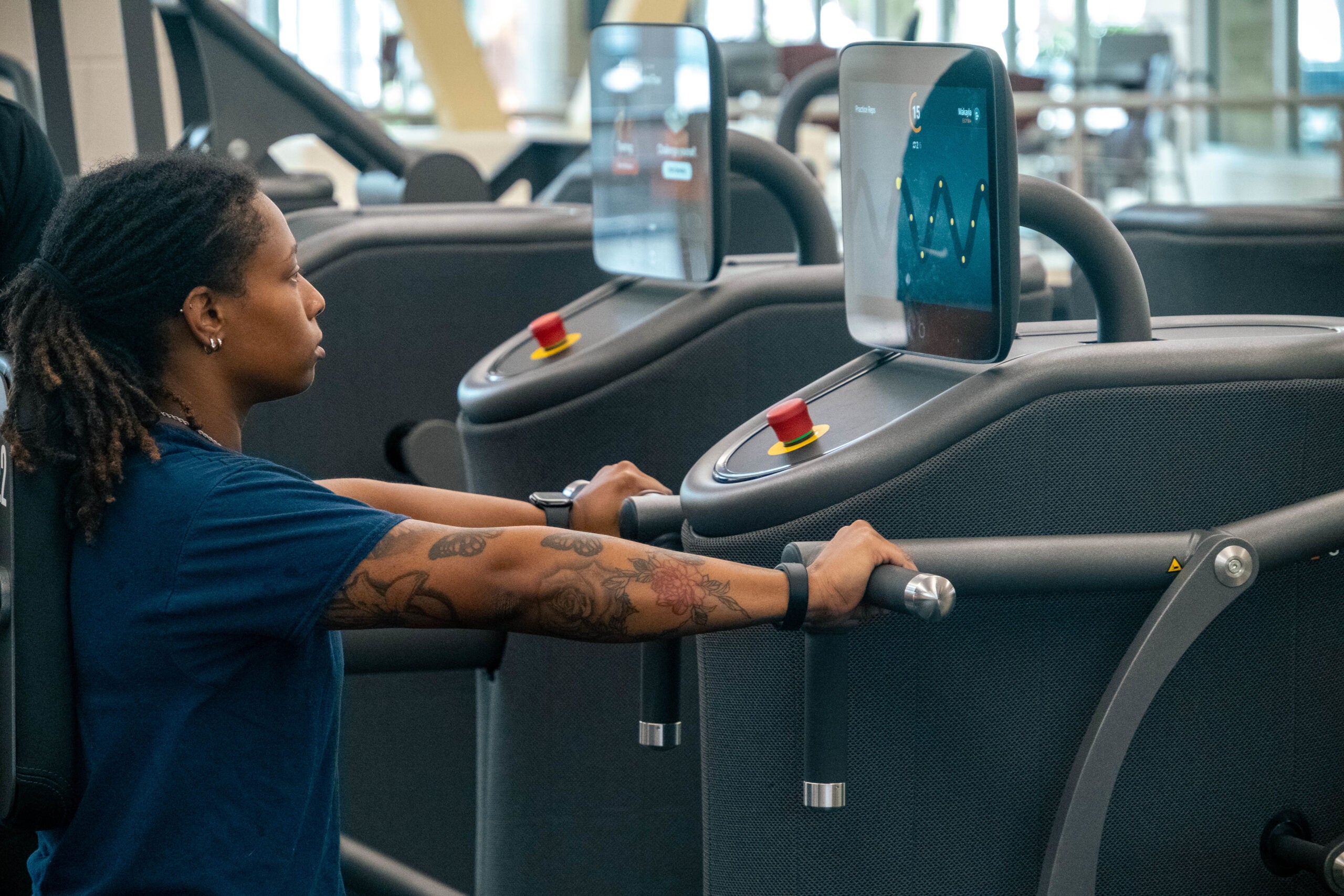 Woman in a blue shirt holds treadmill handles, watching a touchscreen console at a gym machine.