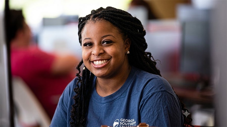 Smiling Black woman with braided hair wearing a blue Georgia Southern University shirt in a bright office setting.