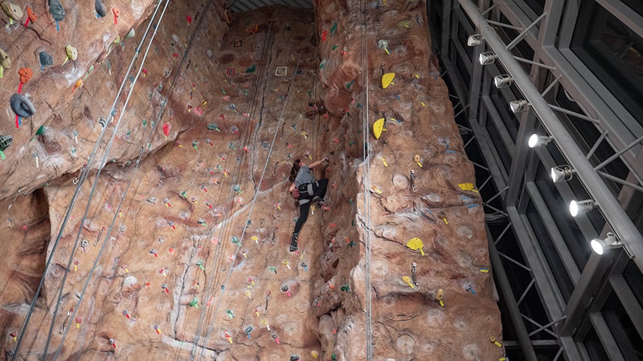A climber in a harness ascends an indoor rock wall with multiple colored holds and safety ropes overhead in a modern gym.