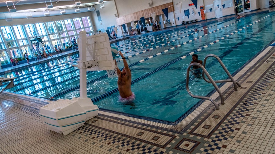 Indoor swimming pool with a player dunking a basketball at a movable backboard by the edge of the pool, lane dividers visible in the water.