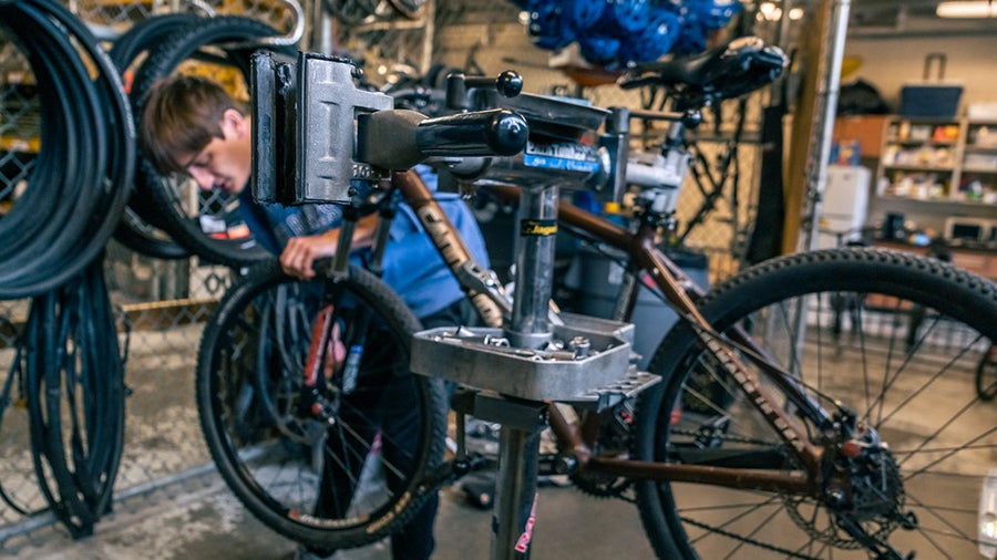 Bicycle mounted on a repair stand as a technician works on its drivetrain in a cluttered workshop.