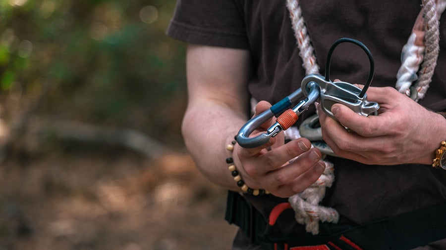 Close-up of a person outdoors holding a carabiner and a climbing harness with rope tails nearby.