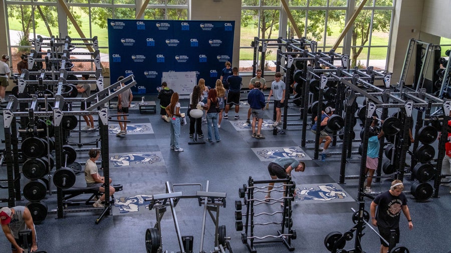 Gym with racks of barbells and weight plates; group of people gathered near a blue backdrop stage at the far end, likely a workshop or presentation.