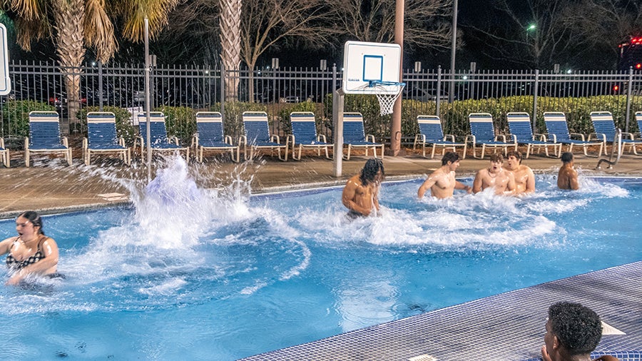 Nighttime outdoor pool scene with several people splashing and playing in the water, surrounded by empty lounge chairs and a basketball hoop in the background.
