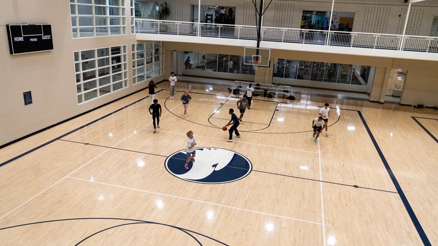 Indoor basketball gym viewed from above with players on the court, a large blue-and-white logo at center, and a balcony running along the second floor.