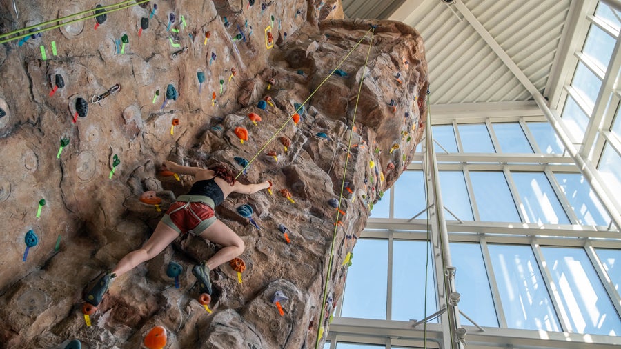 Person rock climbing indoors, wearing a harness and using colorful handholds on a textured wall with a safety rope above.