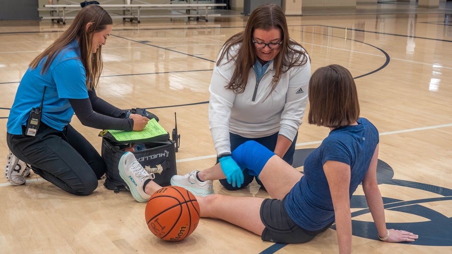 Gym scene: a knee-injured athlete sits on the floor while two trainers assist; one notes on a clipboard, a basketball nearby.