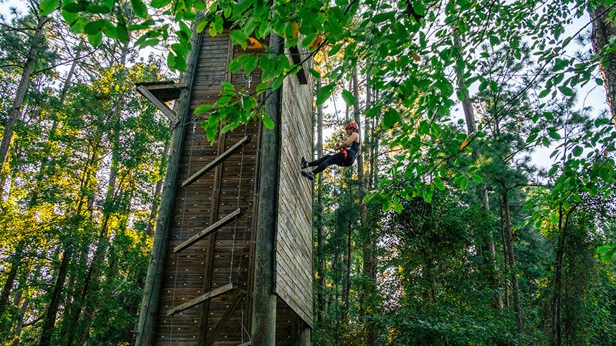 Person wearing a harness climbs a tall wooden outdoor climbing wall in a forested area.