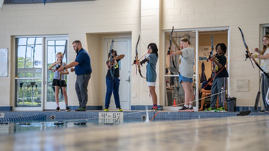 Instructor coaching children in archery along a pool deck inside a recreation center.