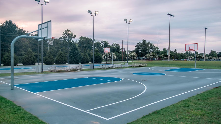 Outdoor basketball courts with blue playing surfaces, white lines, benches, and tall lights at dusk.