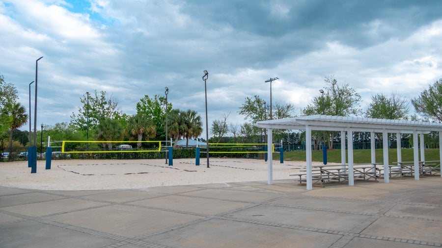 Outdoor public park featuring a sand volleyball court with a yellow net and blue posts, surrounded by trees and light poles, with a covered picnic area and benches to the right.