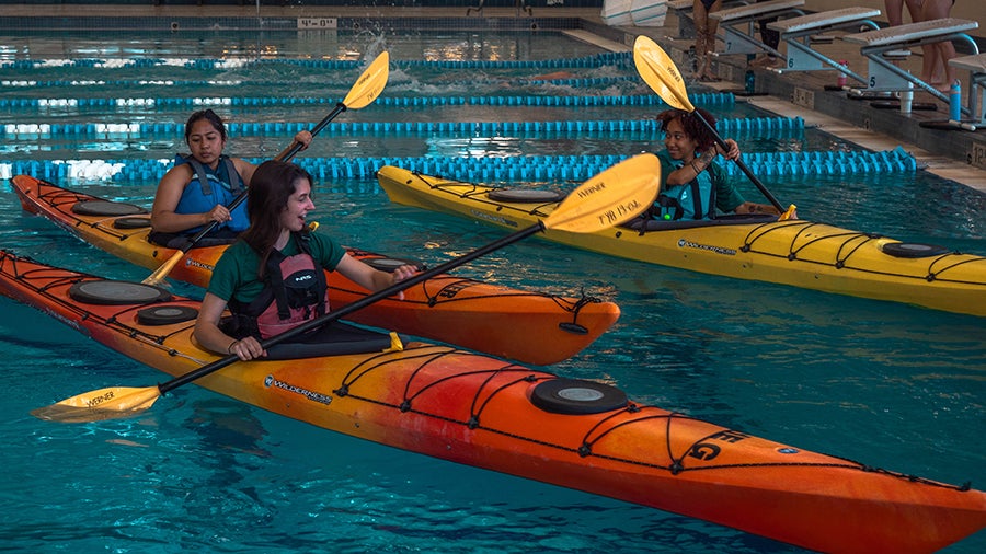 Three people in colorful kayaks paddling in an indoor pool, orange and yellow kayaks visible with lane lines in the water.