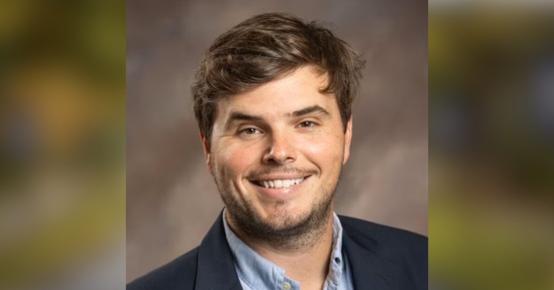 Head-and-shoulders portrait of a smiling man in a navy blazer and light blue shirt, brown hair.