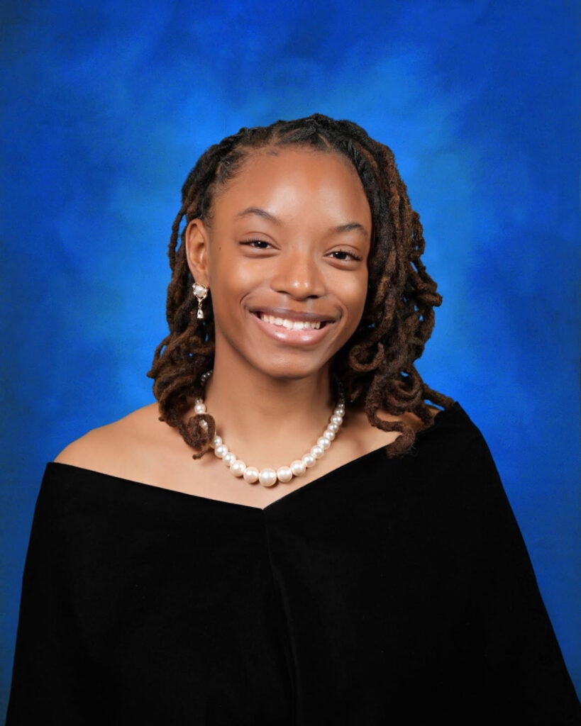 Portrait of a smiling woman, wearing a pearl necklace and earrings against a blue studio background.