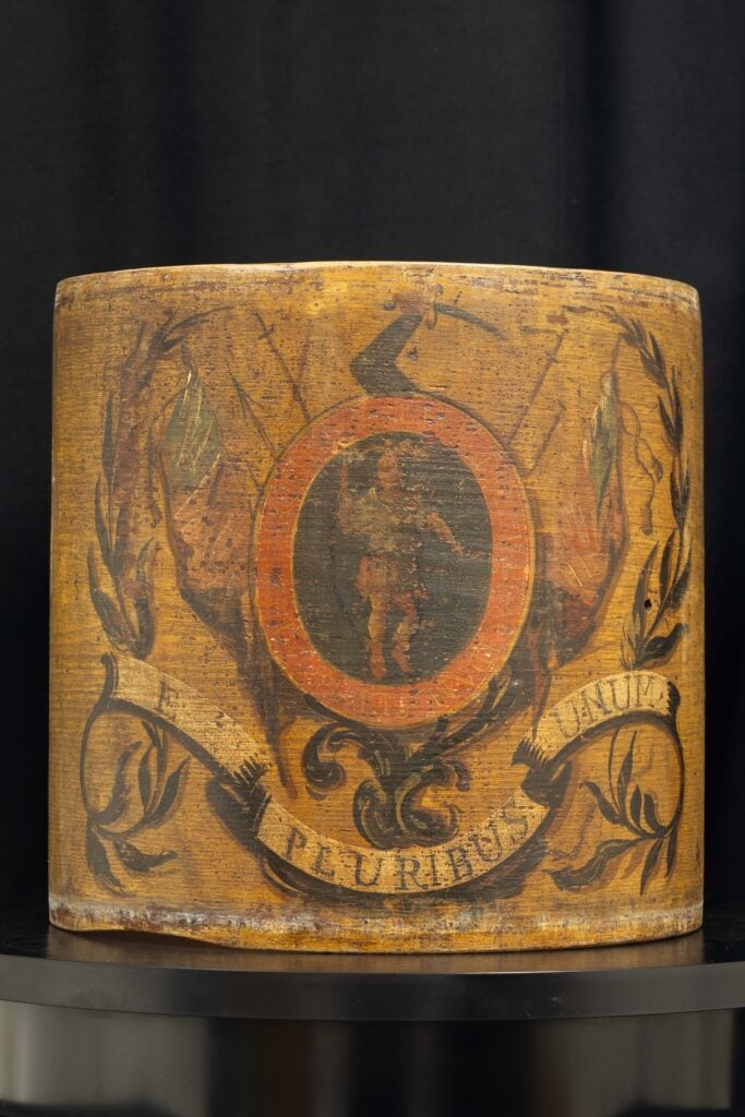 Close-up of a weathered Revolutionary War drum with a red circular emblem and faded decorations including a coat of arms. There is a human figure in the center of the red oval emblem on the tan, worn drum.