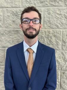 Man wearing a blue suit, white shirt, and tan tie stands against a textured stone wall, wearing glasses.