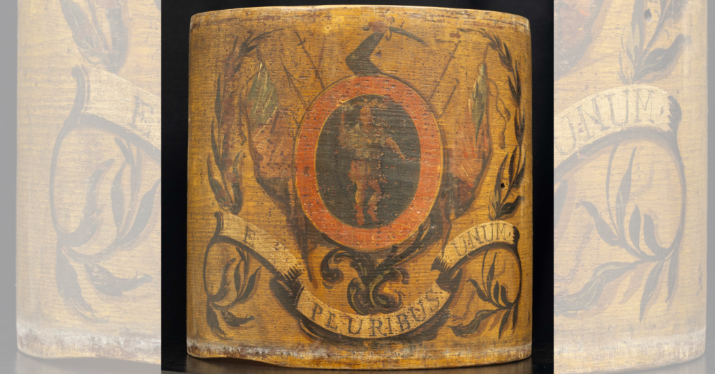 Close-up of a weathered Revolutionary War drum with a red circular emblem and faded decorations including a coat of arms. There is a human figure in the center of the red oval emblem on the tan, worn drum.