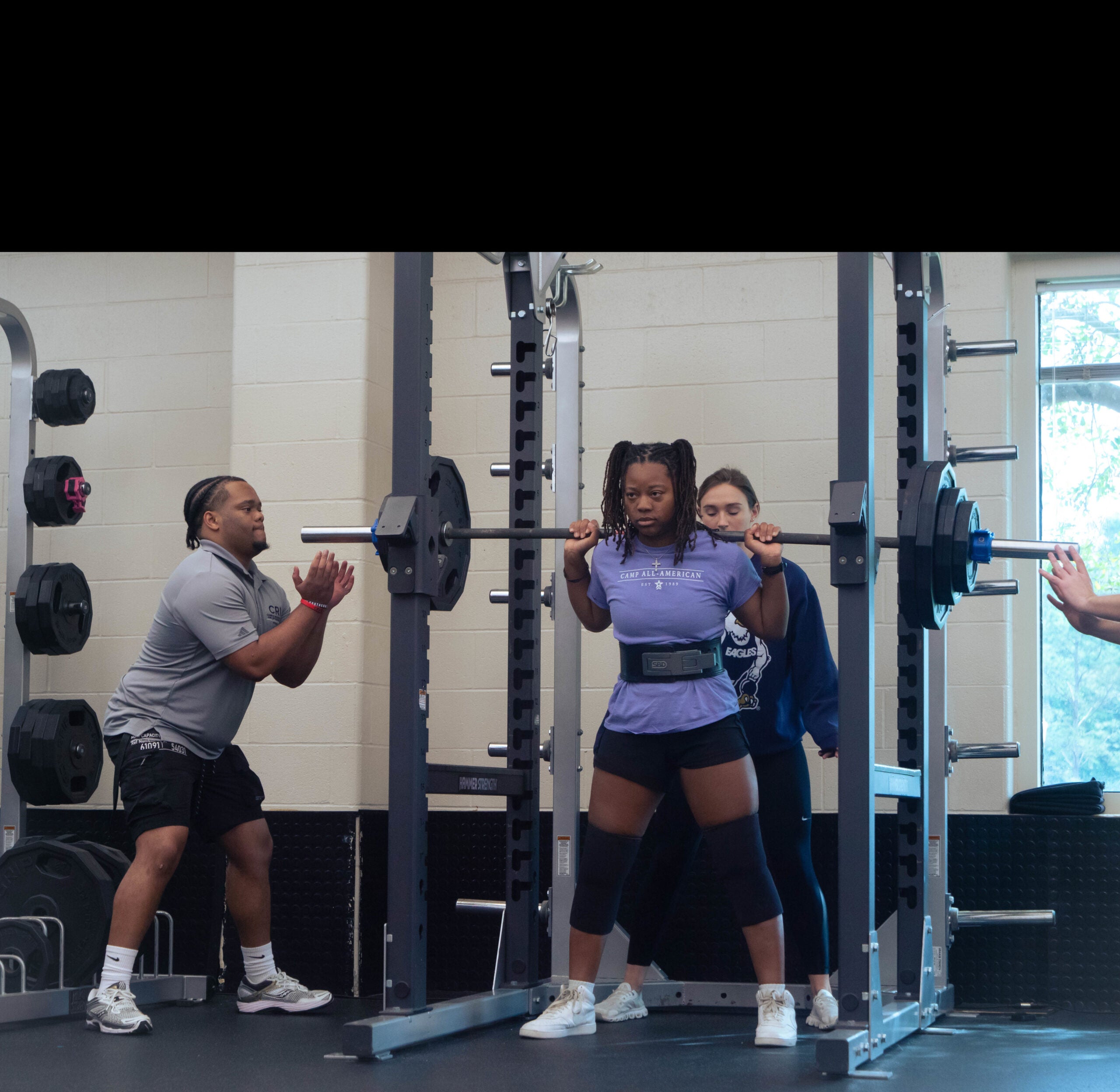People in a gym spotting a woman doing barbell squats at a squat rack; others stand nearby for support.