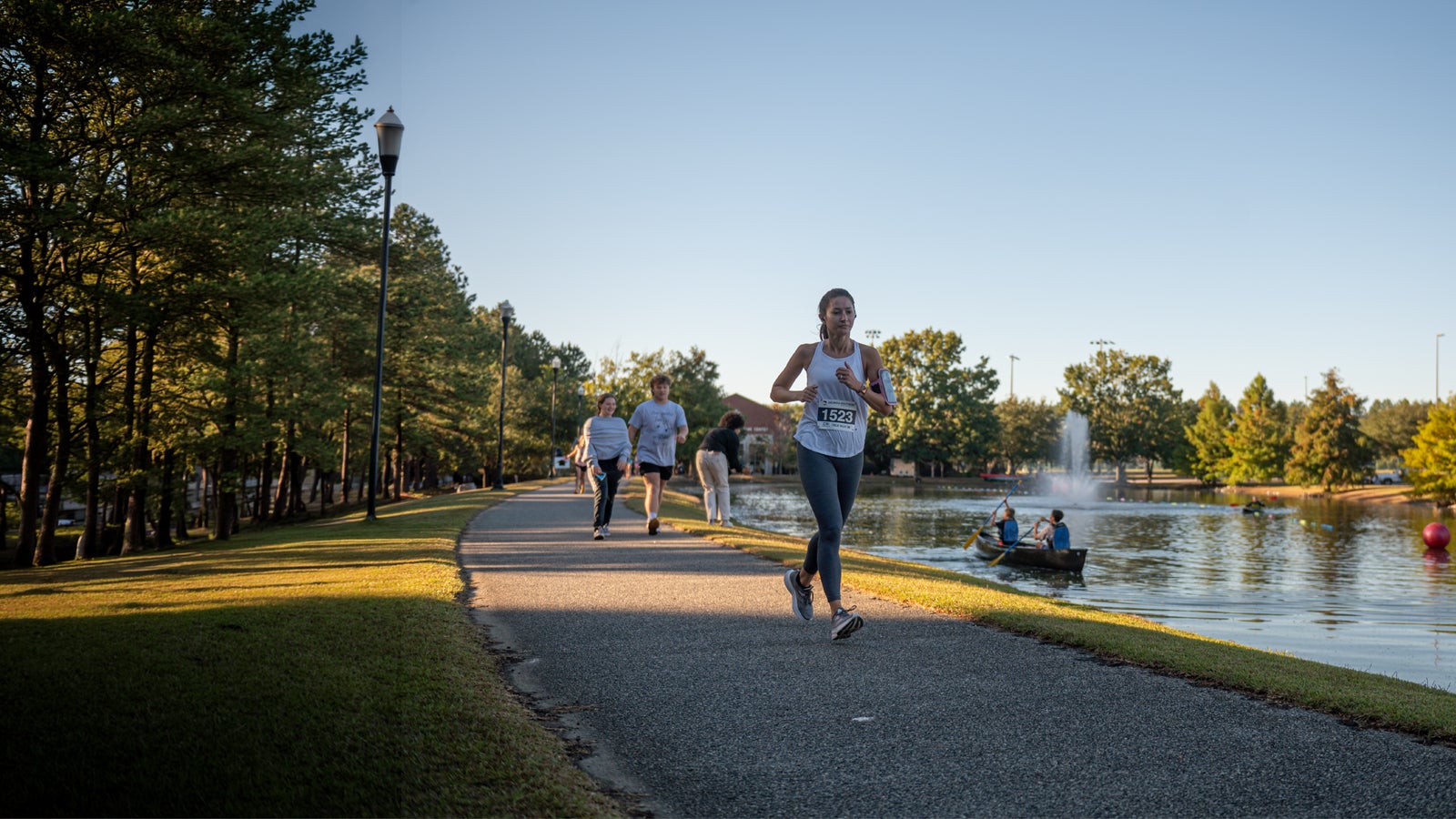 Female runner with bib 1523 jogging along a lakeside park path; others walk in the background as rowboats glide on the water and a fountain shoots up in the lake.