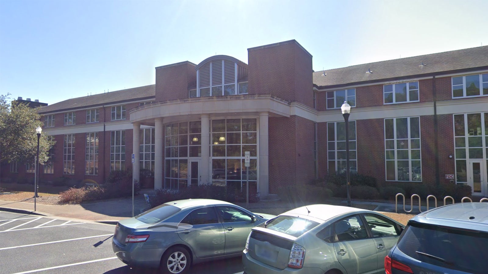 Brick institutional building with a large glass entrance and curved front, cars parked in the foreground on a sunny day.