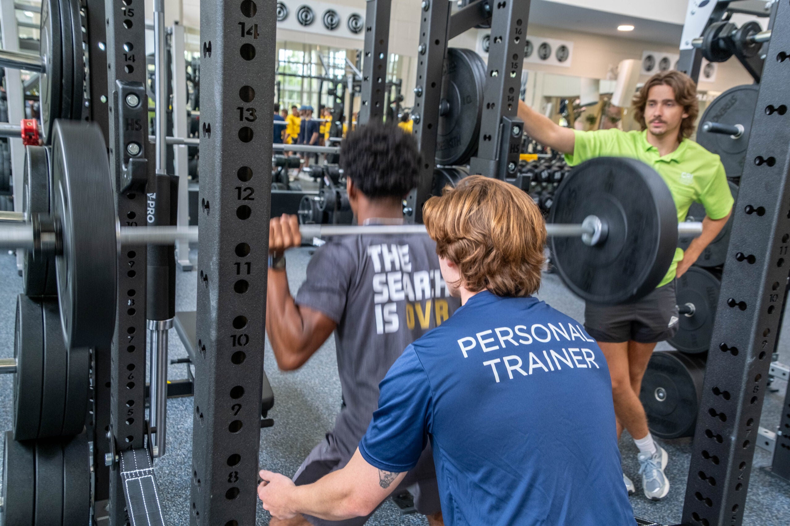 Lifters in a gym squat rack: a person in blue with 'PERSONAL TRAINER' on the back bench-pressing, another lifter nearby, and a trainer in a bright green shirt spotting.