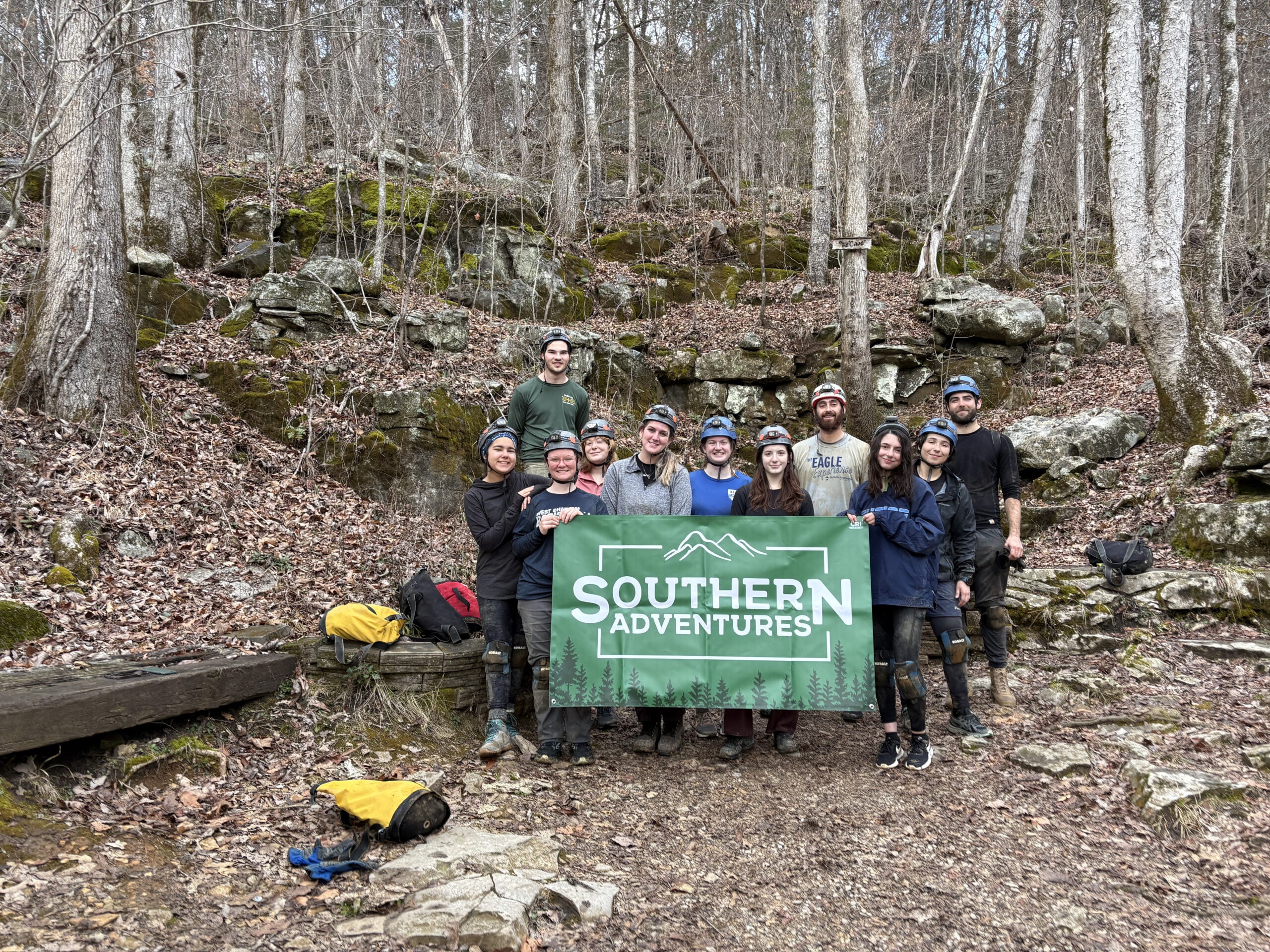 Group of people wearing helmets posing with a green Southern Adventures banner in a rocky, forested area with fallen leaves and mossy boulders.