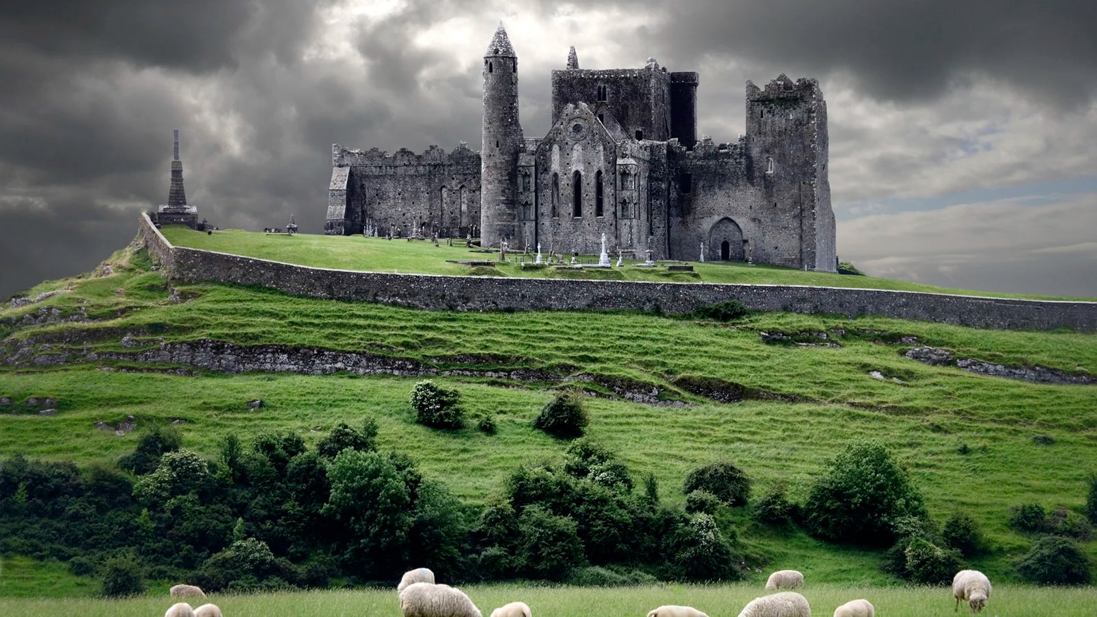 Ruined medieval castle atop a grassy hill with sheep in the foreground under dark storm clouds.