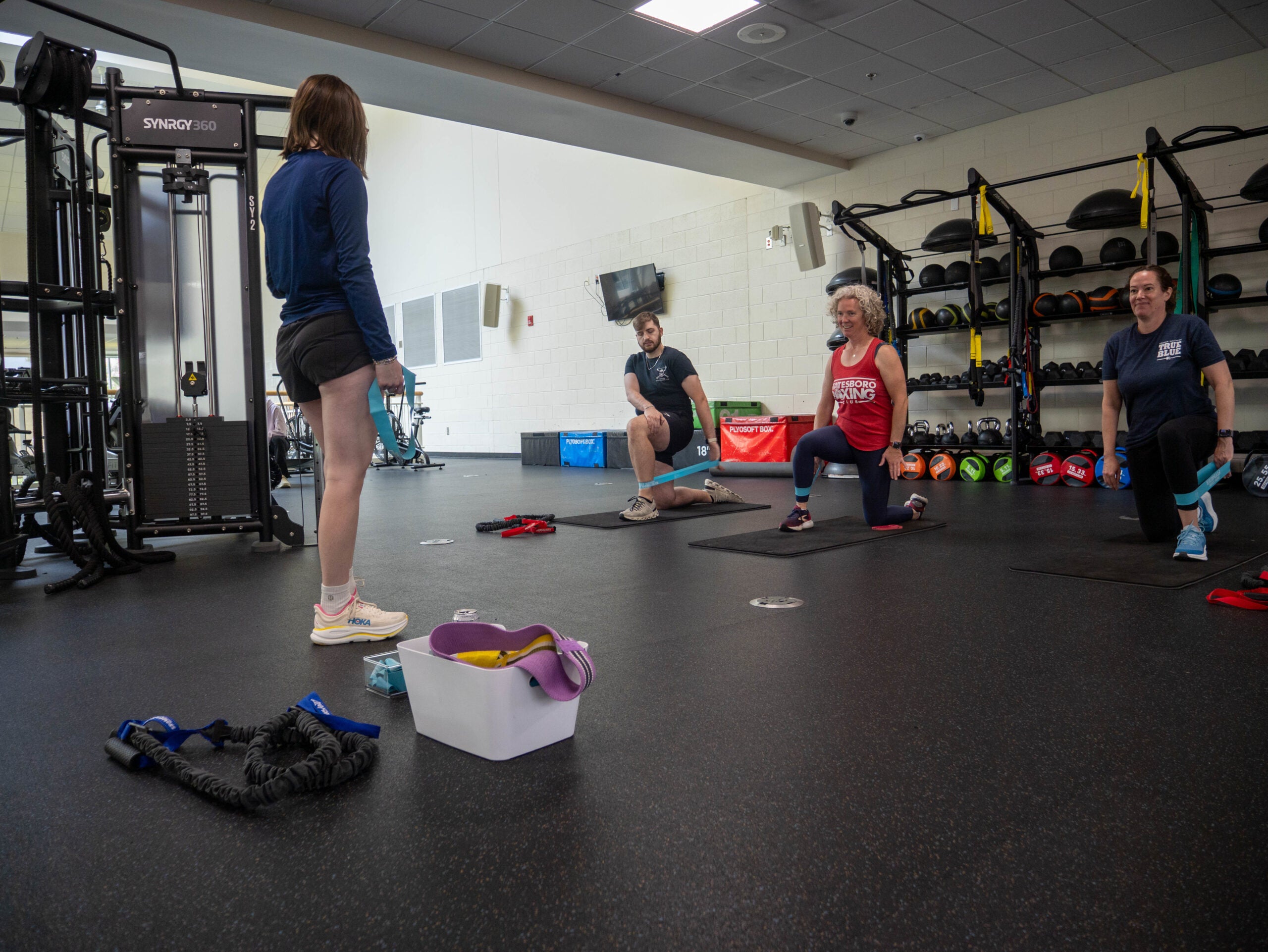 Gym scene with participants kneeling on mats, using resistance bands during a group workout; weights and shelves in the background.
