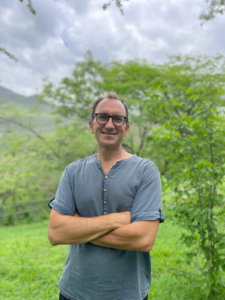 Smiling man with glasses standing outdoors in a green, wooded field with arms crossed.