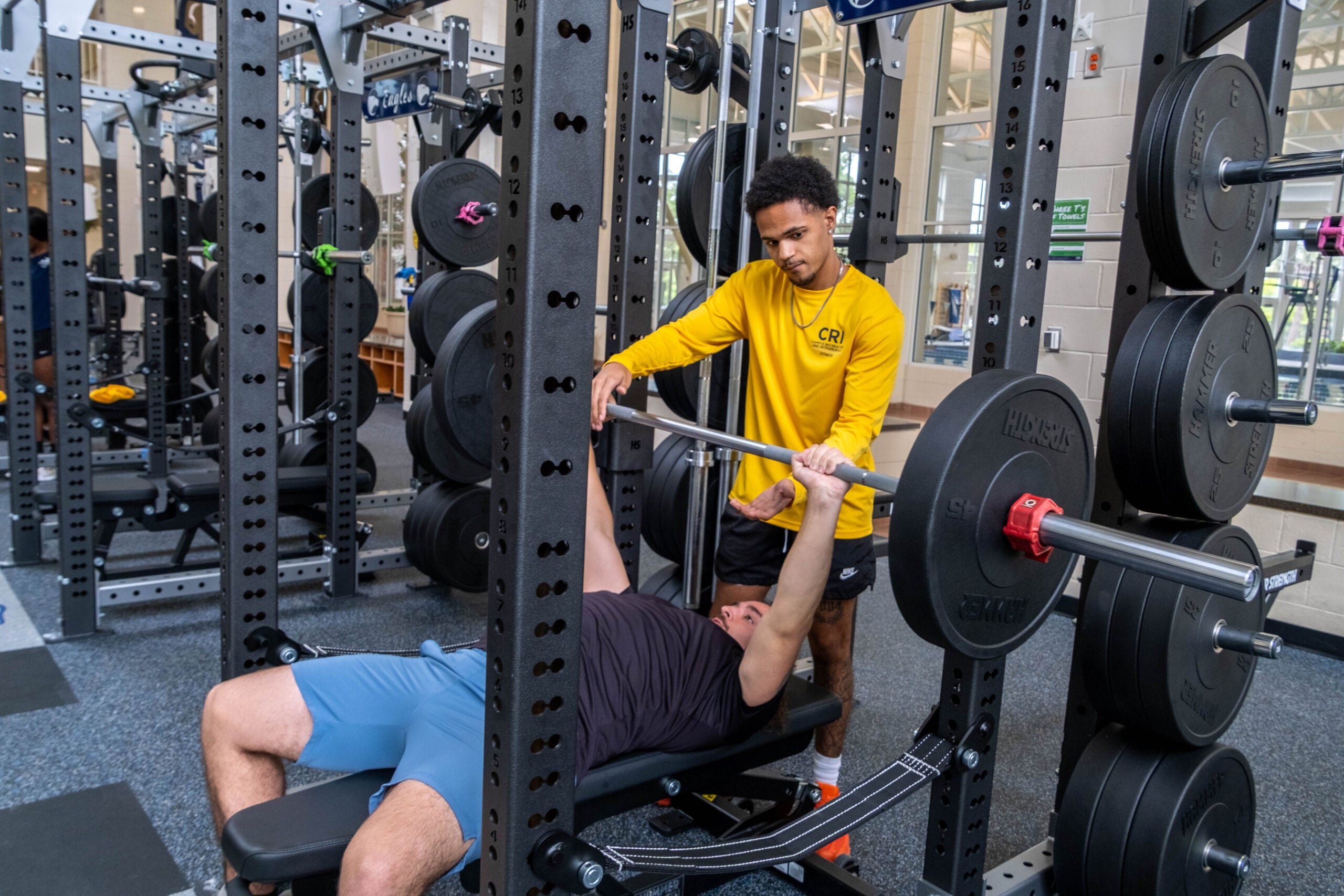 Gym scene with a trainer in a yellow shirt assisting a lifter during a bench press on a loaded rack with plates nearby.