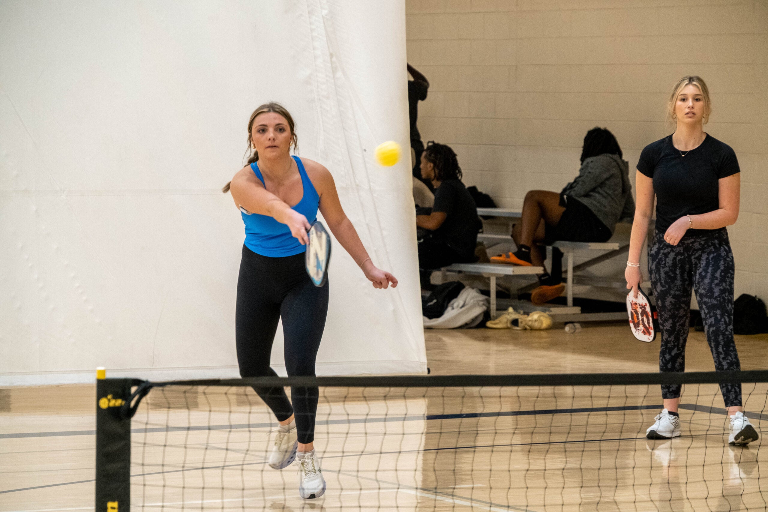 Two women playing indoor paddle tennis or pickleball near a net; one in a blue tank hits a yellow ball toward the net while the other stands with a paddle watching.