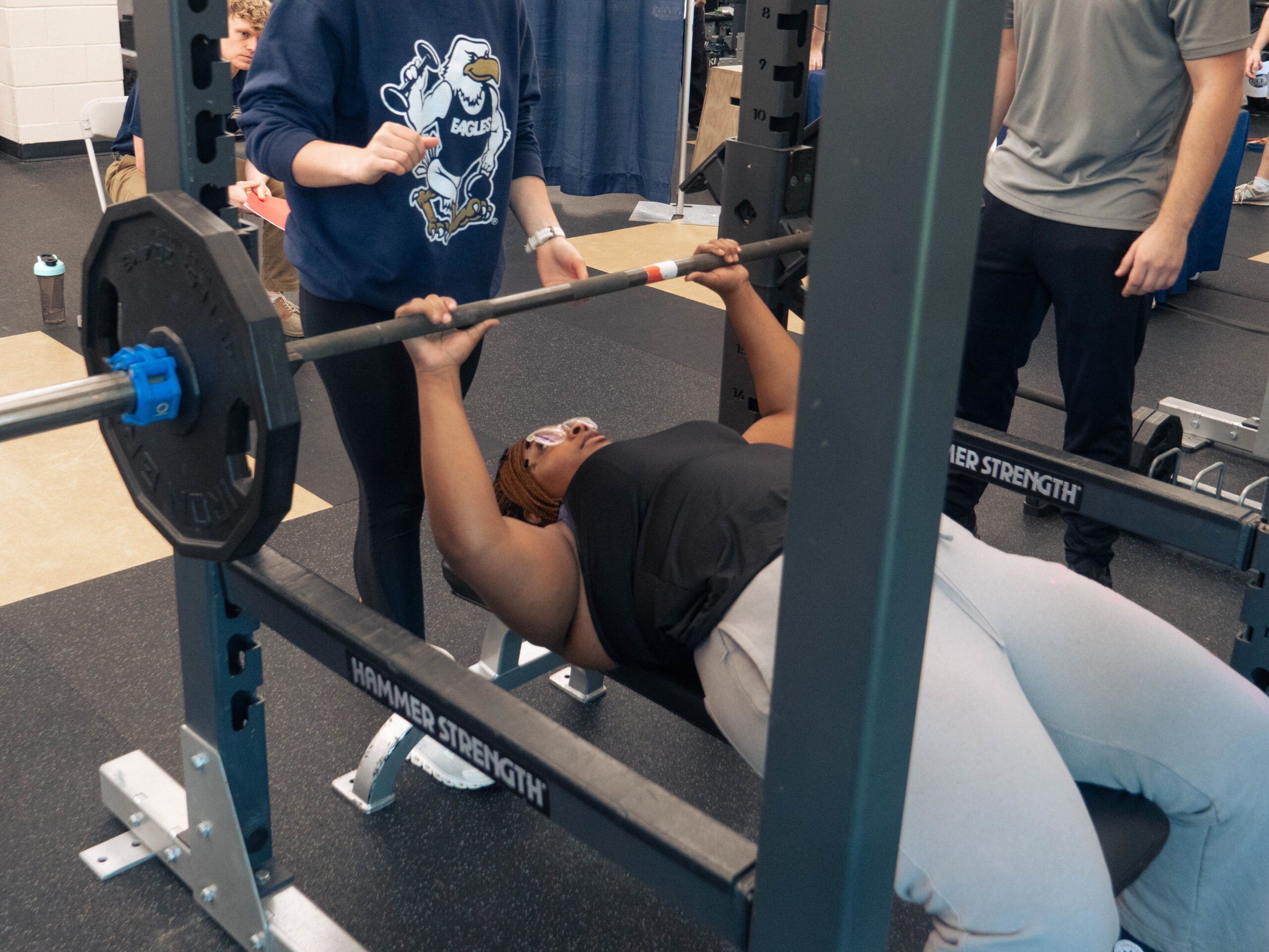 Person lying on a bench press, lifting a barbell while a spotter stands behind them in a gym setting (weight room)