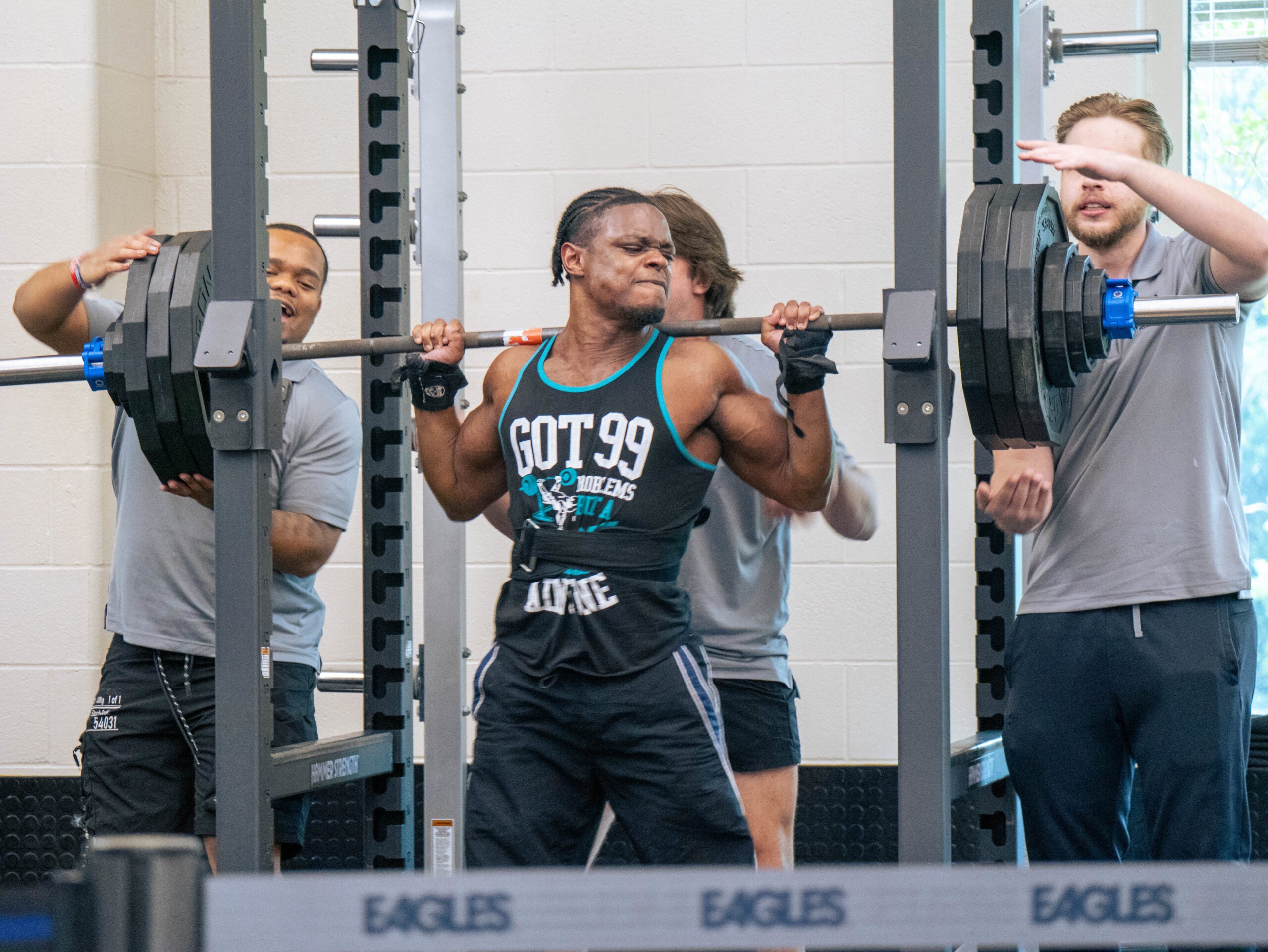 Lifters in a gym: man in a weightlifting singlet struggles under a heavy barbell while two spotters support from the sides in a squat rack.
