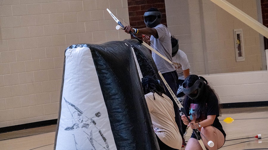 Group of students in a gym wearing safety goggles, using long sticks to hit a small white ball toward a padded obstacle.