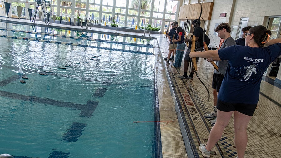 Group of people at a poolside holding bows, practicing archery indoors beside the water.