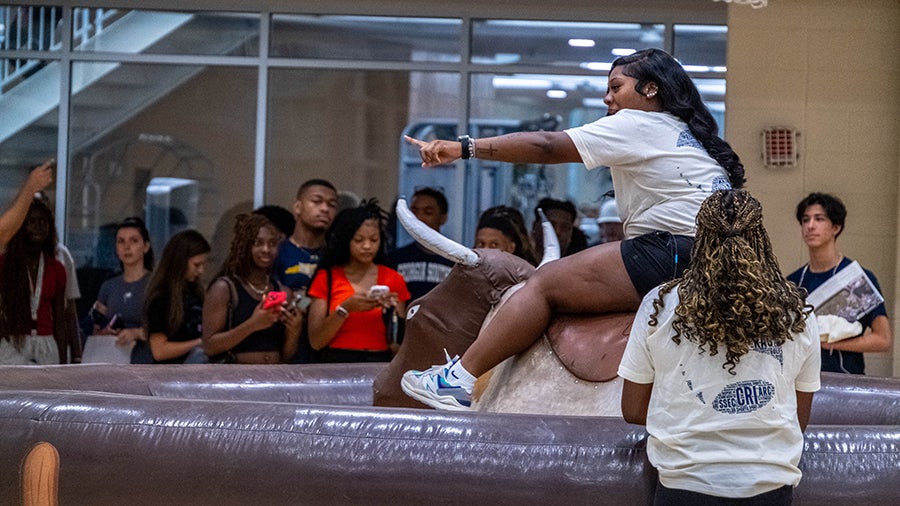 Young woman in a white shirt rides a mechanical bull indoors, while a helper stands nearby and a crowd of students watches, some recording on phones.