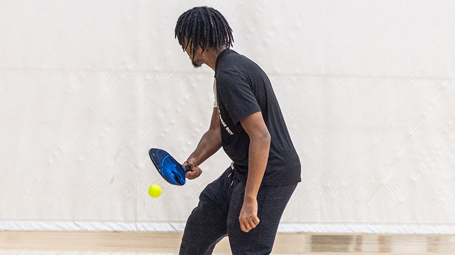 Man in a black shirt playing paddle ball indoors, swinging a blue paddle at a bright yellow ball nearby.