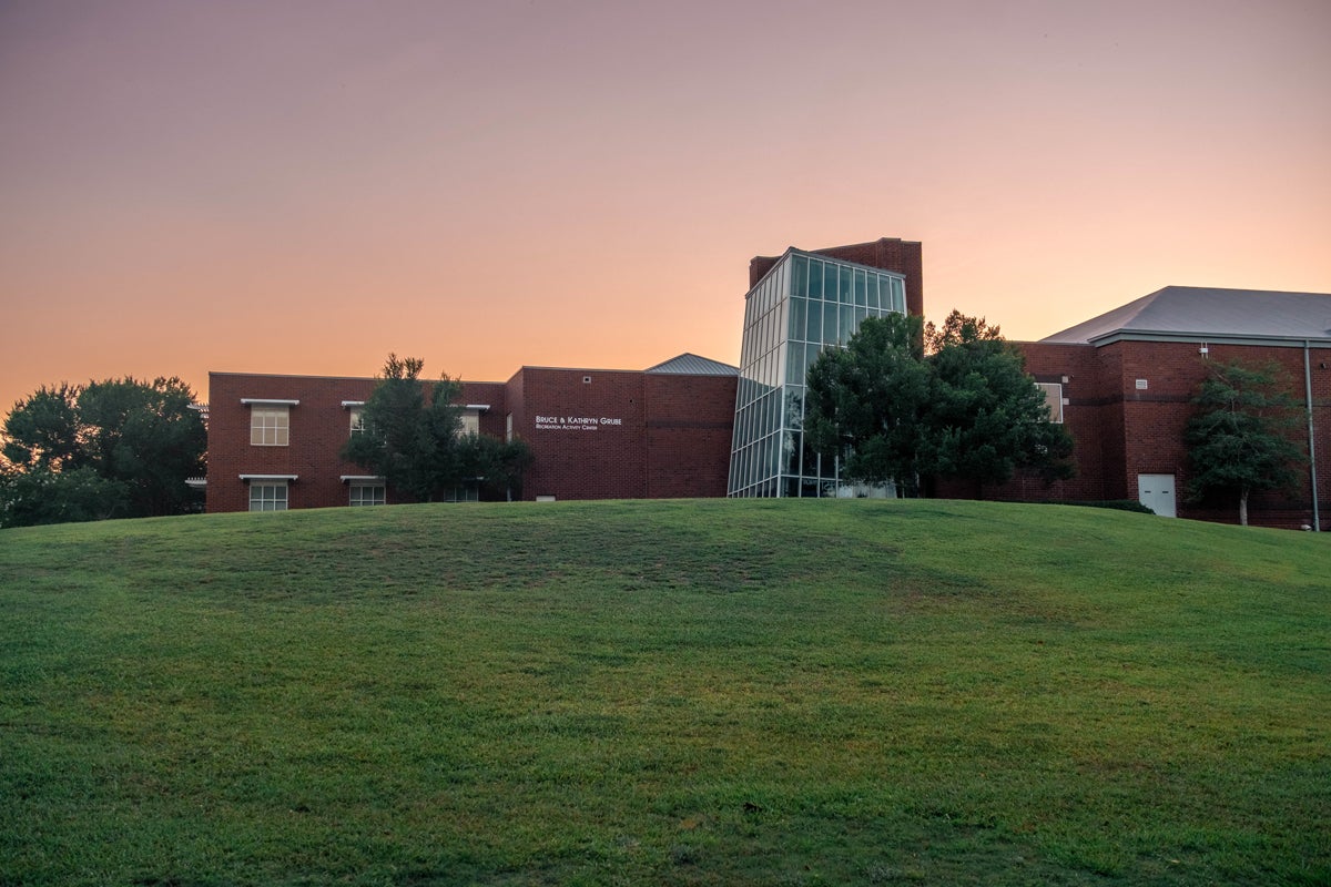 Brick campus building with a glass-paneled central atrium on a grassy hill at sunset.