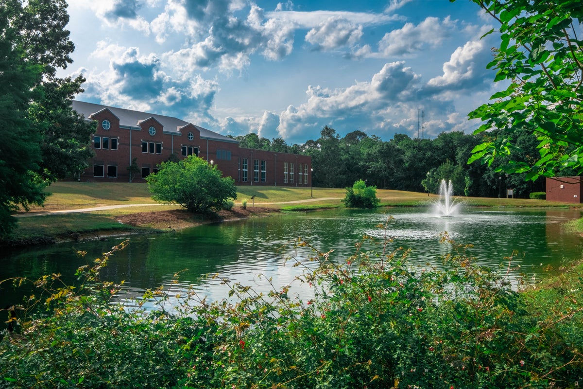Campus pond with a fountain, a brick collegiate building in the background, and green landscaping under a partly cloudy blue sky.