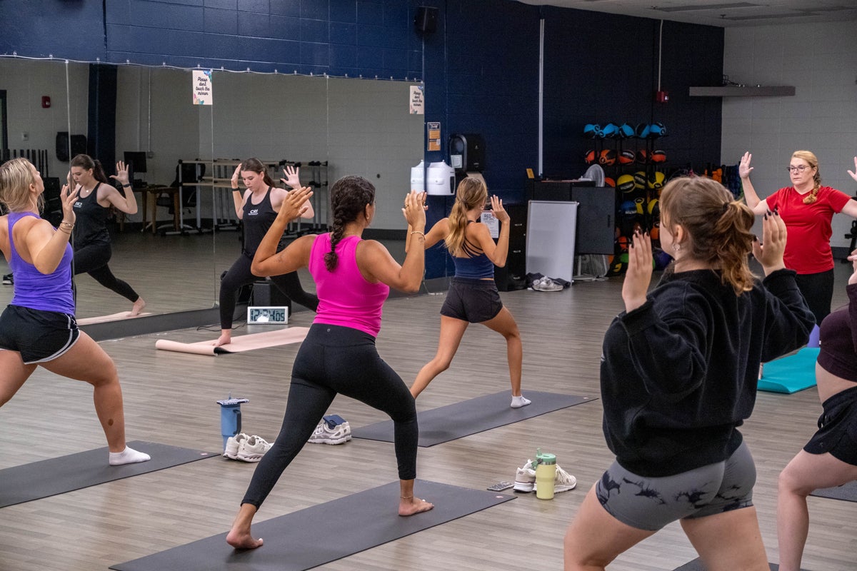 Group fitness class in a gym studio, participants on yoga mats raising hands in a pose during a workout.