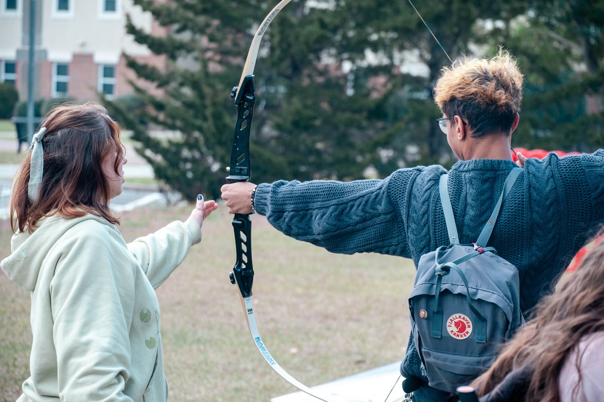 Two people outdoors practicing archery: one holds a recurve bow while the other extends a hand toward the string, nearby grassy field background