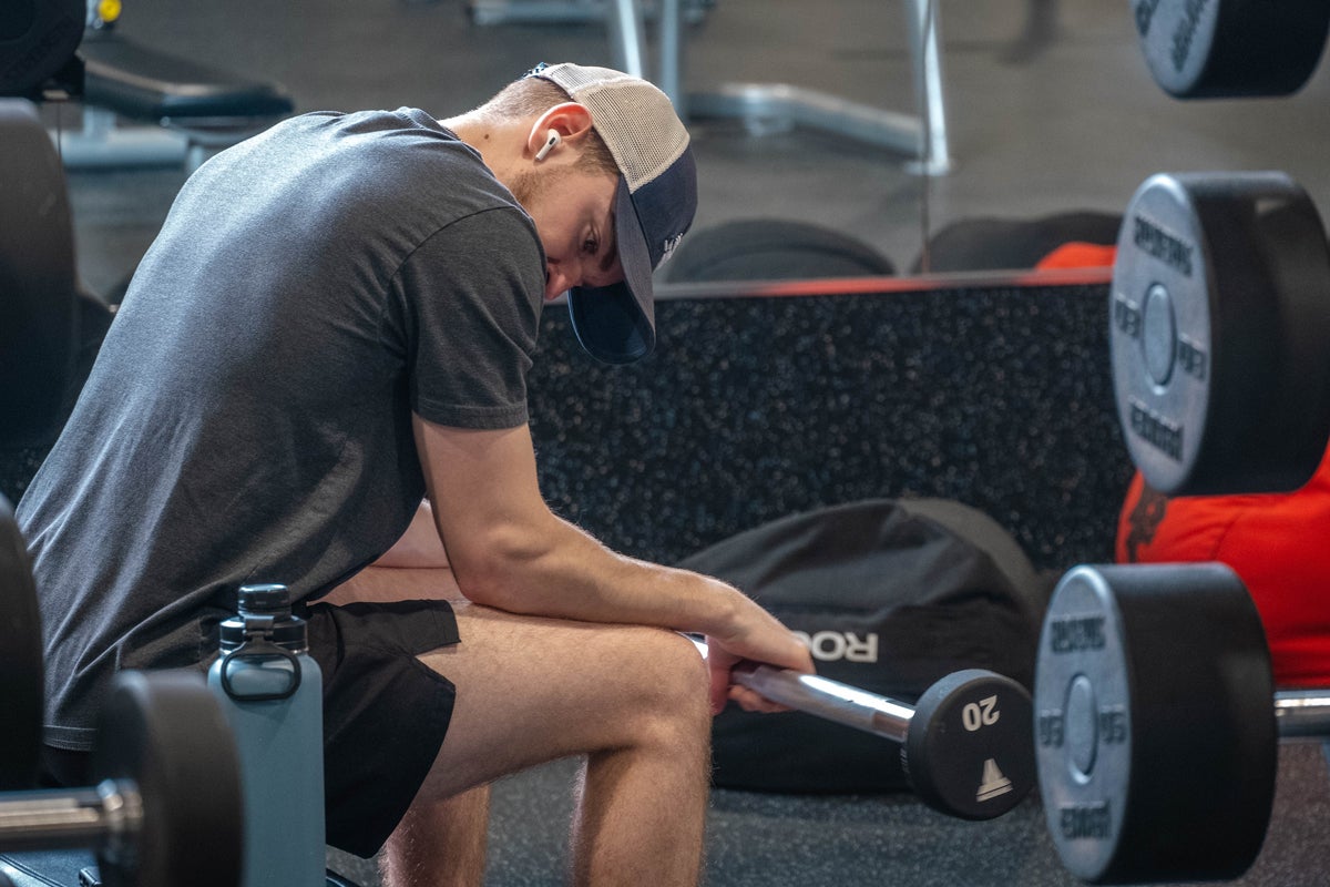 Man in a gray shirt and cap sits on a bench in the gym, gripping a short barbell with 20 lb weight plates nearby, wearing wireless earbuds.