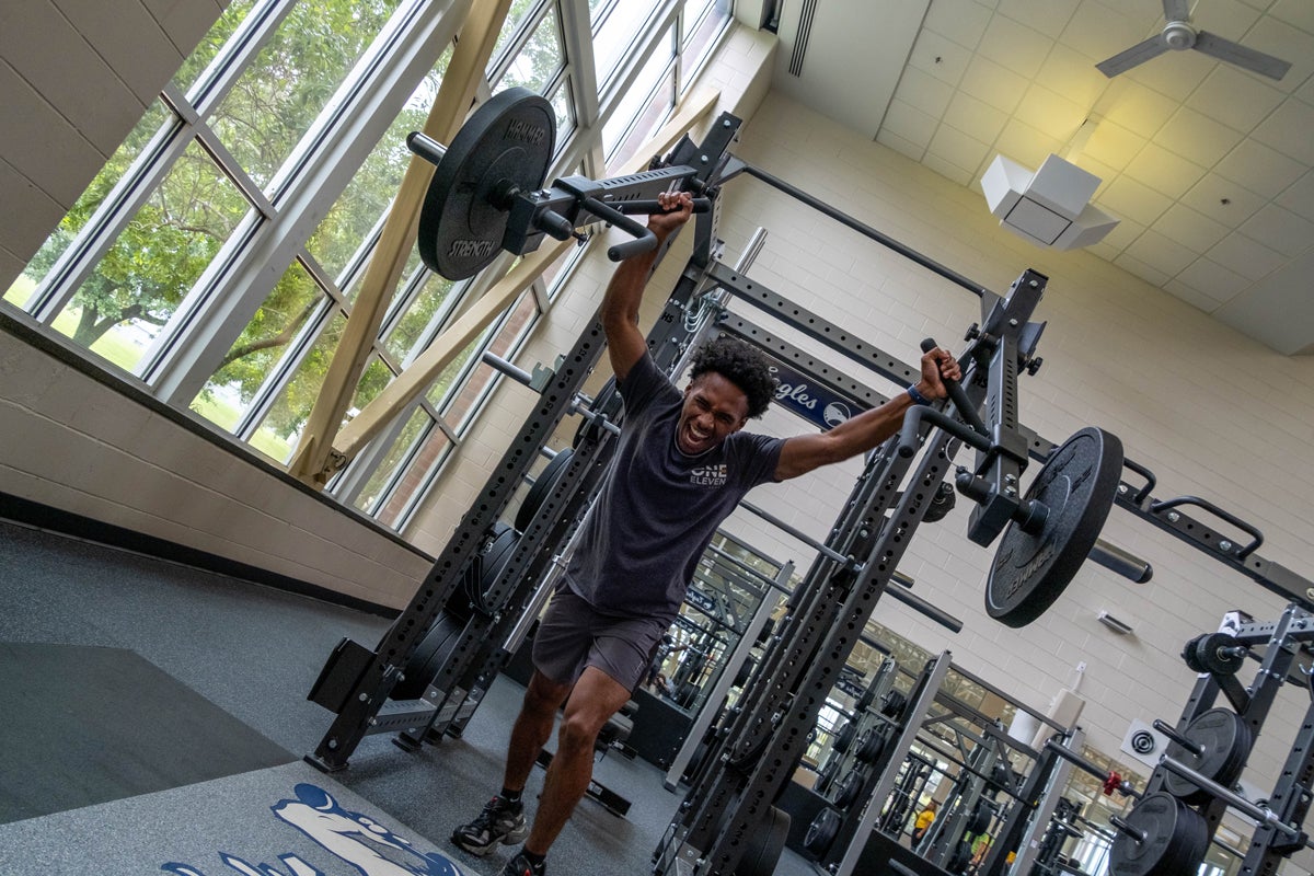 Man lifting a heavy barbell overhead inside a power rack at a bright gym.