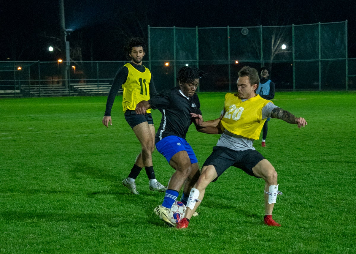 Two players in yellow bibs and one in black contest the ball during a night soccer match.
