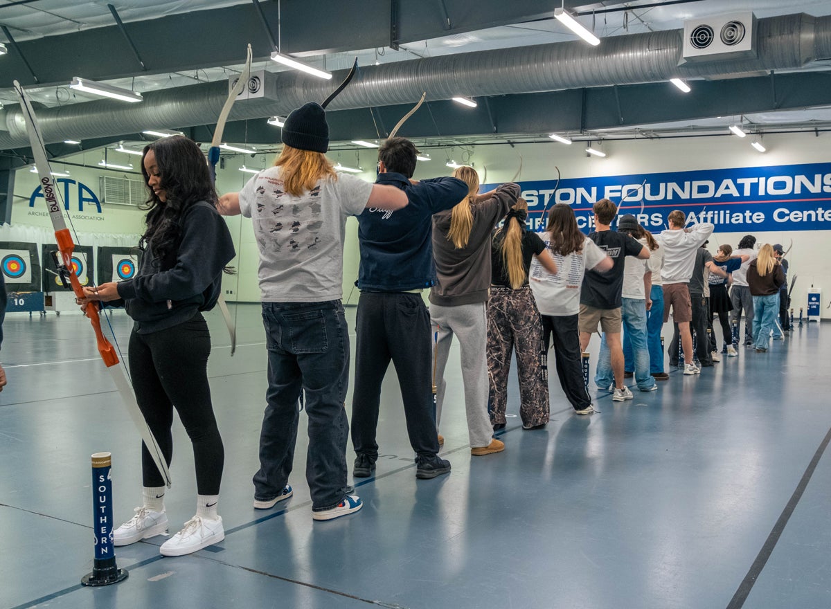 Line of people in an indoor archery range, each archer drawing a bow, targets visible at the back of the room.