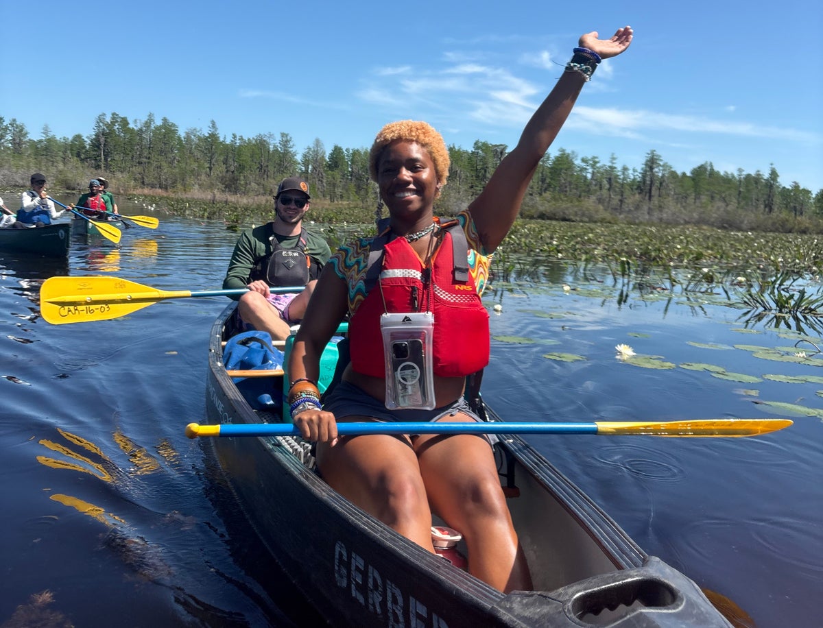 Smiling woman in a red life jacket sits in the front of a canoe, her left arm raised in the air as teammates paddle on a sunny lake with lily pads nearby.