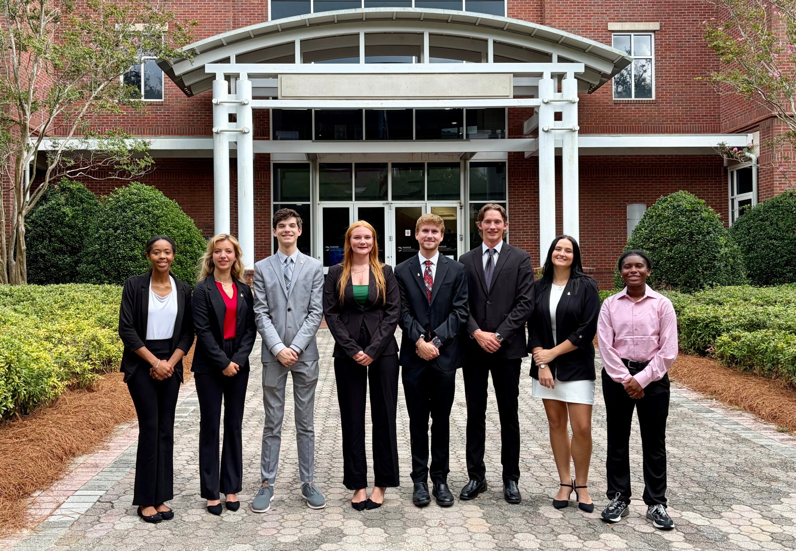Group of eight students in formal attire posing outdoors in front of a brick building with glass doors and landscaping behind them.